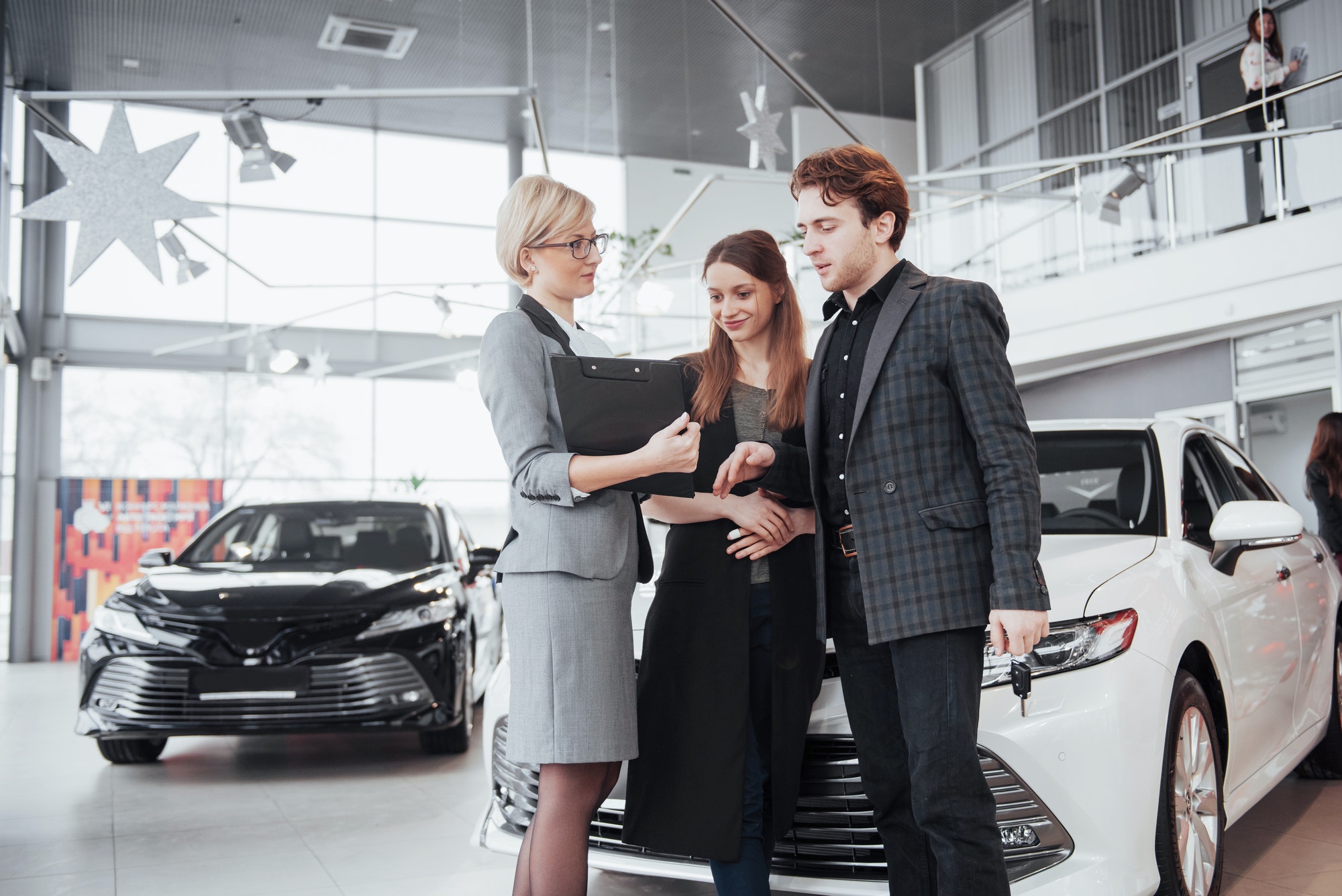 Gorgeous loving couple posing together near their new car at the car dealership showroom salon